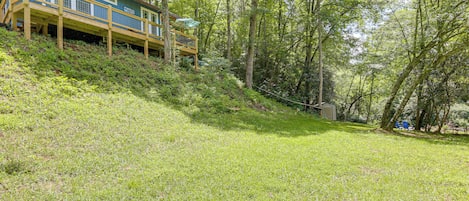A house with a wooden deck and a covered area, surrounded by a grassy slope and trees.
