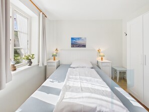 Ett sovrum med en säng, sängbord, ett fönster med gardiner, en tavla som hänger på väggen och ett litet bord.