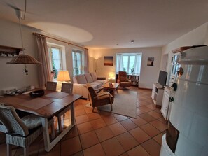 A living room with a sofa, armchairs, a coffee table, a dining table with chairs, a TV, and a refrigerator. - Ingke Vacation Rental with a Fireplace – Family Vacations on Föhr - Boldixumer Straße (Wyk auf Föhr)