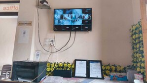Een receptiebalie met een flatscreenmonitor waarop meerdere camerabeelden te zien zijn, een printer en ingelijste documenten.