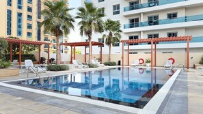 Piscina com espreguiçadeiras e um pergolado, cercada por palmeiras e edifícios modernos.