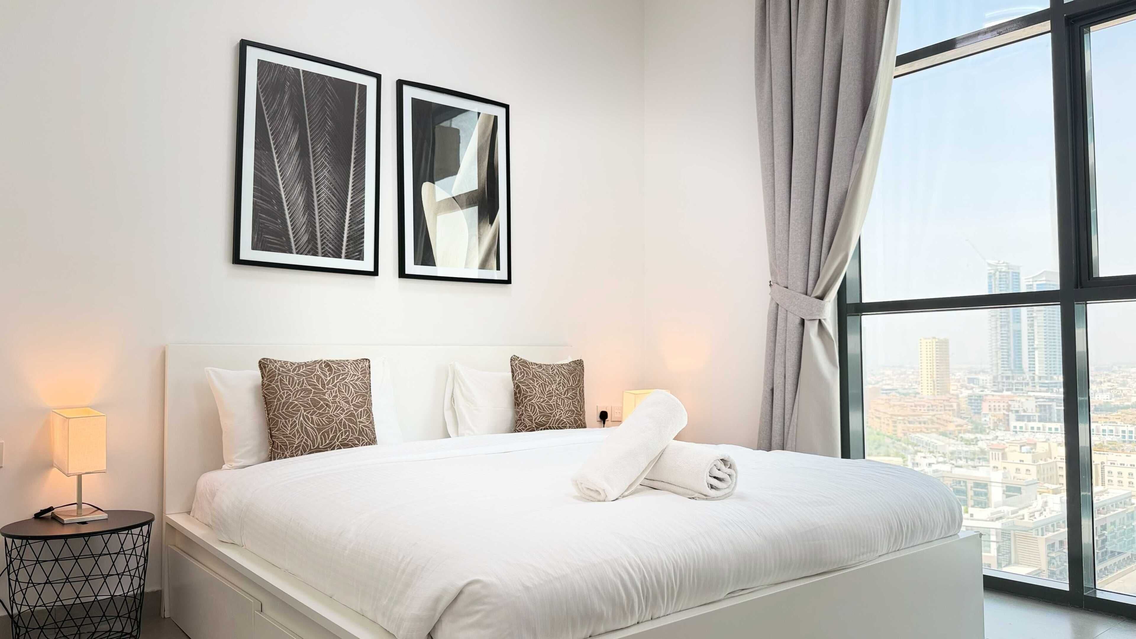 Une chambre d’hôtel avec un grand lit, deux œuvres d’art encadrées au-dessus, une vue sur la ville par une grande fenêtre et une table de chevet avec une lampe.
