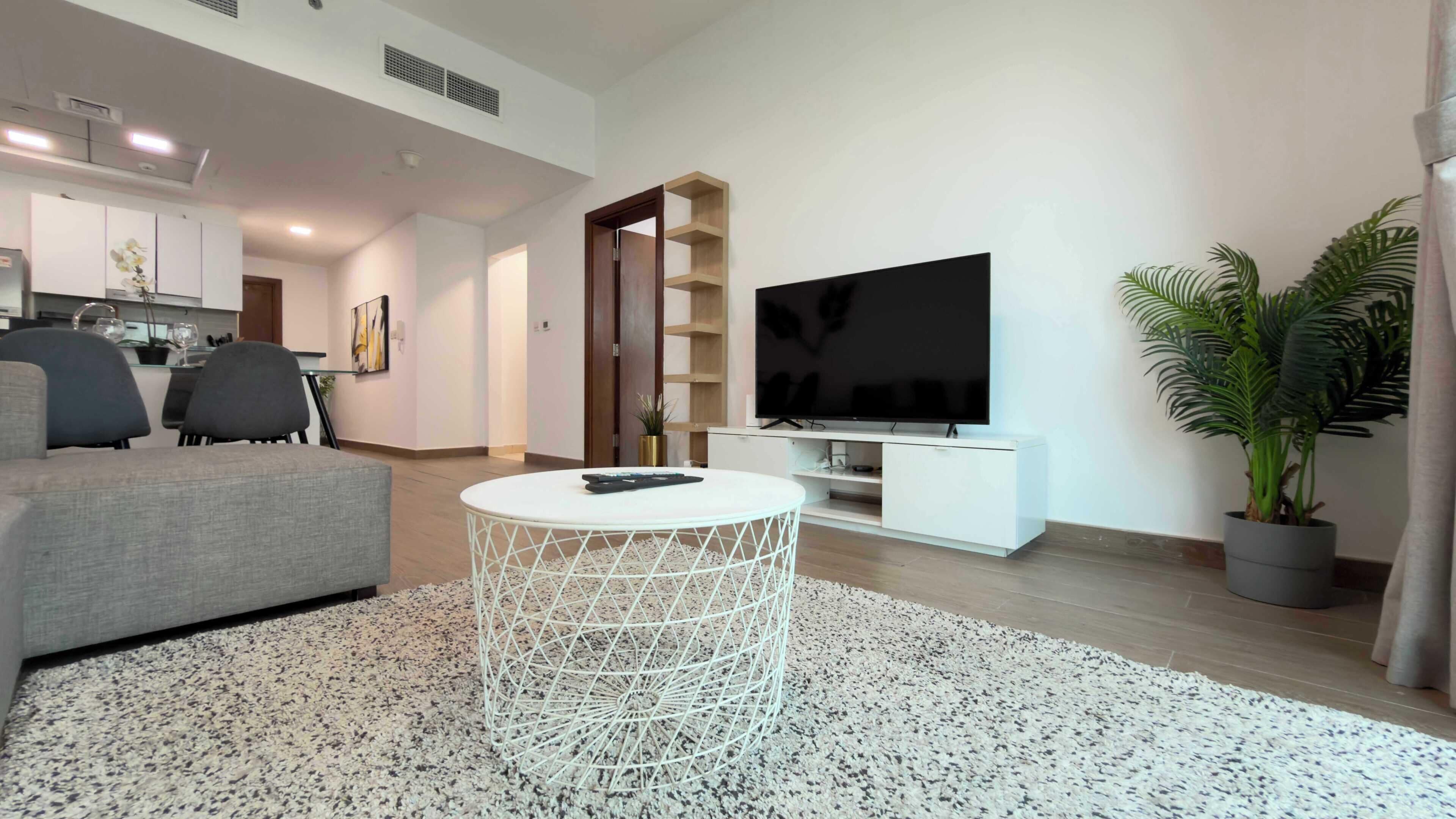 A modern living room with a grey sofa, a white coffee table, a flat-screen TV on a white stand, a kitchen in the background, and a potted plant.