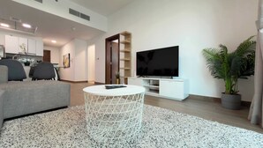 Ein modernes Wohnzimmer mit einem grauen Sofa, einem weißen Couchtisch, einem Flachbildfernseher auf einem weißen Schrank, einer Küche im Hintergrund und einer Topfpflanze.