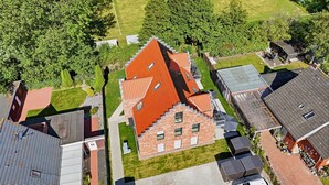 Et boligområde med et stort hus, et markant rødt tag, en velplejet græsplæne og en fodboldbane i baggrunden.