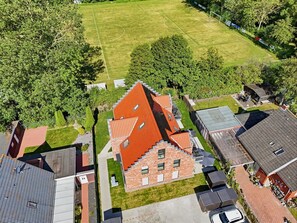 A residential area with a large house featuring a prominent red roof, a well-maintained lawn, and a soccer field in the background. - Küstenkoje - Küstenkoje: New vacation rental with a garden in Norddeich (Norden)