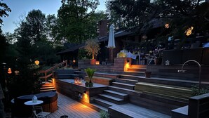 Una terraza de madera con asientos integrados y una pequeña mesa, rodeada de plantas en macetas y una escalera que lleva a un nivel superior.