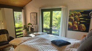 Un dormitorio con una cama grande, una ventana con vistas a zonas verdes, un cuadro en la pared y una planta en maceta en la mesita de noche.