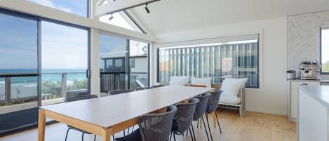 Een moderne keuken met een groot eiland, een eethoek met een lange tafel en stoelen, en uitzicht op de oceaan door grote ramen.