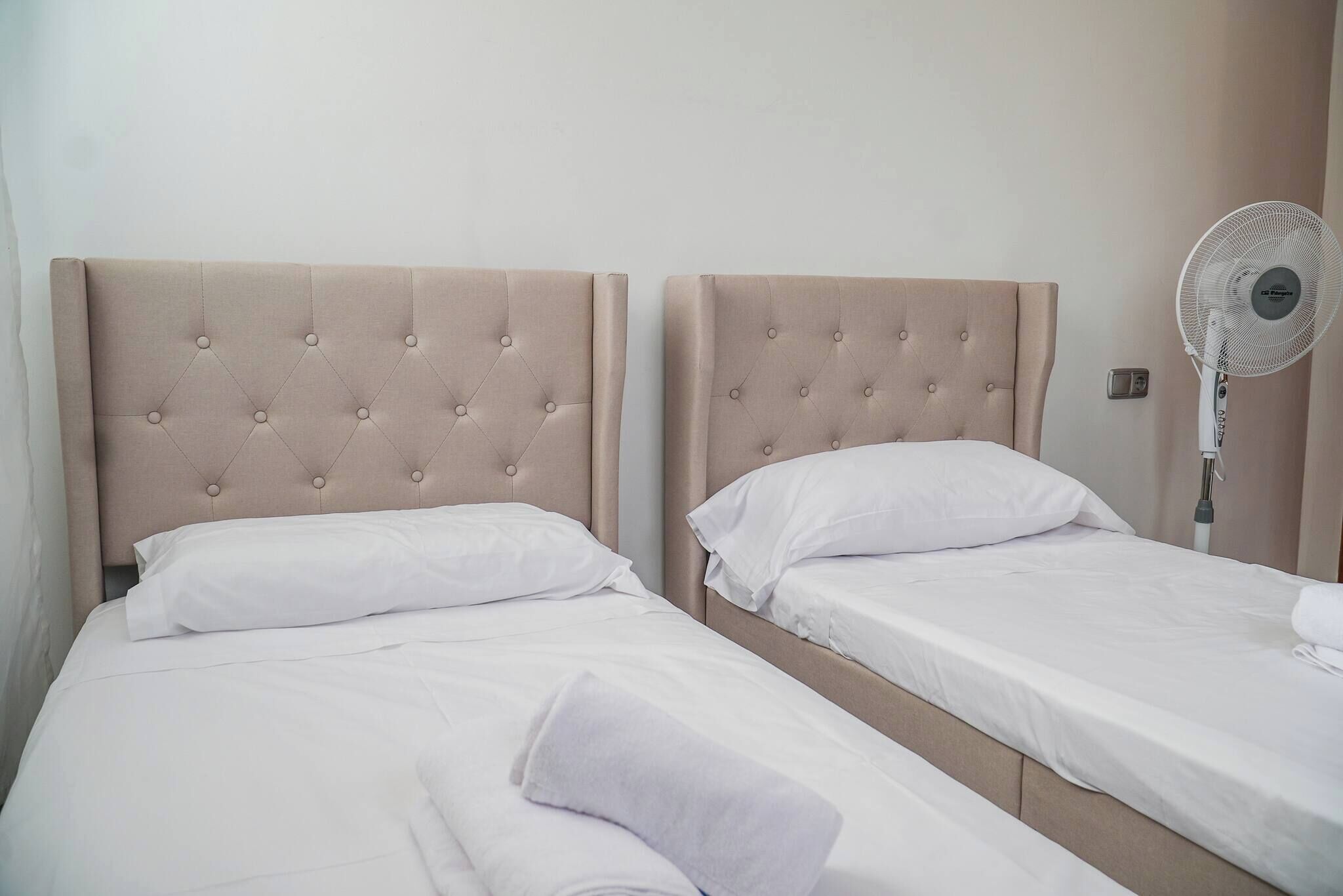 Dos camas individuales con cabeceras, ropa de cama blanca y un ventilador de pie.