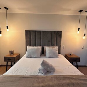 Ein ordentlich bezogenes Bett mit grauem Kopfteil, zwei Nachttische mit Lampen und ein gefaltetes Handtuch auf dem Bett.