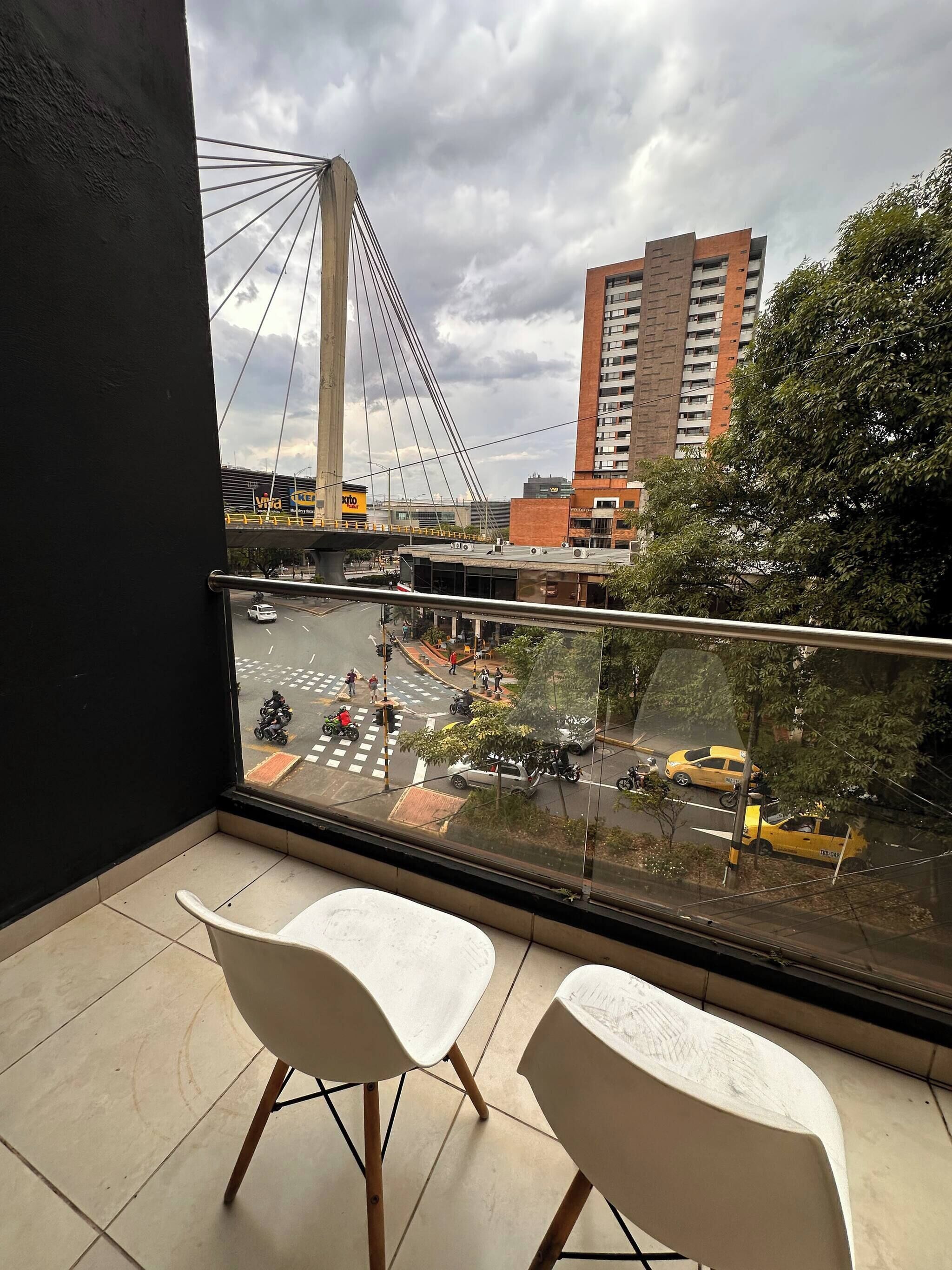 A balcony with two white chairs, a view of a busy street with vehicles and pedestrians, and a large bridge in the background.