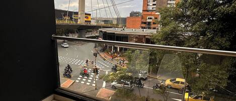 A balcony with two white chairs, a view of a busy street with vehicles and pedestrians, and a large bridge in the background.