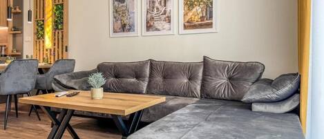 A modern living room with a grey sectional sofa, a wooden coffee table, and a dining area with grey chairs. There are three framed artworks on the wall.