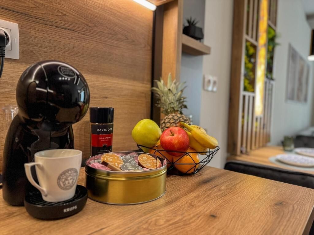 Una taula de fusta amb una cafetera, una tassa, una llauna de galetes i una cistella de fruita que conté pomes, plàtans i una pinya.
