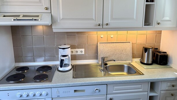 A kitchen with white cabinets, a stainless steel sink, a coffee maker, and a stove. - Reiherweg, Apartment 4 - Beautiful 2-room vacation apartment with a south-facing balcony, quiet and (Sylt)