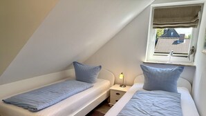A small, triangular attic bedroom with two single beds, a nightstand with a lamp, and a window with a view of a house. - Reiherweg, Apartment 4 - Beautiful 2-room vacation apartment with a south-facing balcony, quiet and (Sylt)