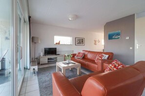 A modern living room with a sofa set, a coffee table, a TV, and a window with a view of the outdoors. - Coastal Bungalow with Private Garden (Makkum)