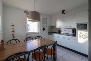 A compact kitchen with white cabinets, a wooden dining table, and a hanging light fixture. - Coastal Bungalow with Private Garden (Makkum)