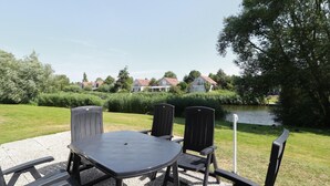 An outdoor seating area with a table and chairs, a gravel surface, and a view of a lake with houses in the background. - Waterfront Makkum Bungalow with Jetty (Makkum)