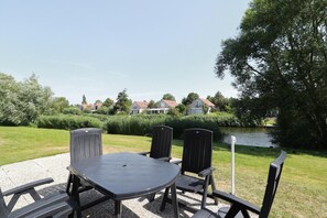 An outdoor seating area with a table and chairs, a gravel surface, and a view of a lake with houses in the background. - Waterfront Makkum Bungalow with Jetty (Makkum)