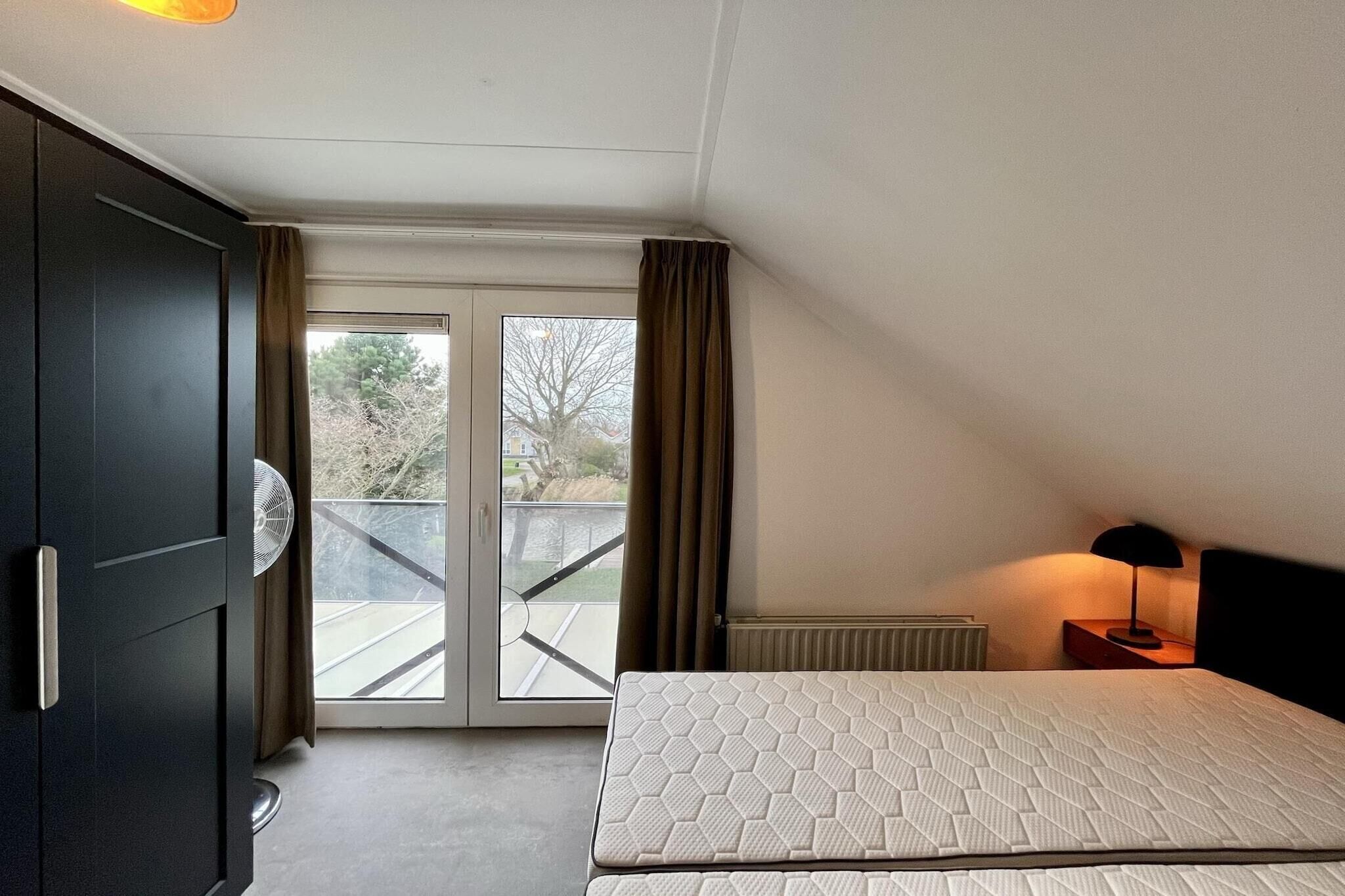 A bedroom with a bed, a radiator, a nightstand with a lamp, a window with curtains, and a view of a balcony.