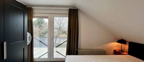 A bedroom with a bed, a radiator, a nightstand with a lamp, a window with curtains, and a view of a balcony.