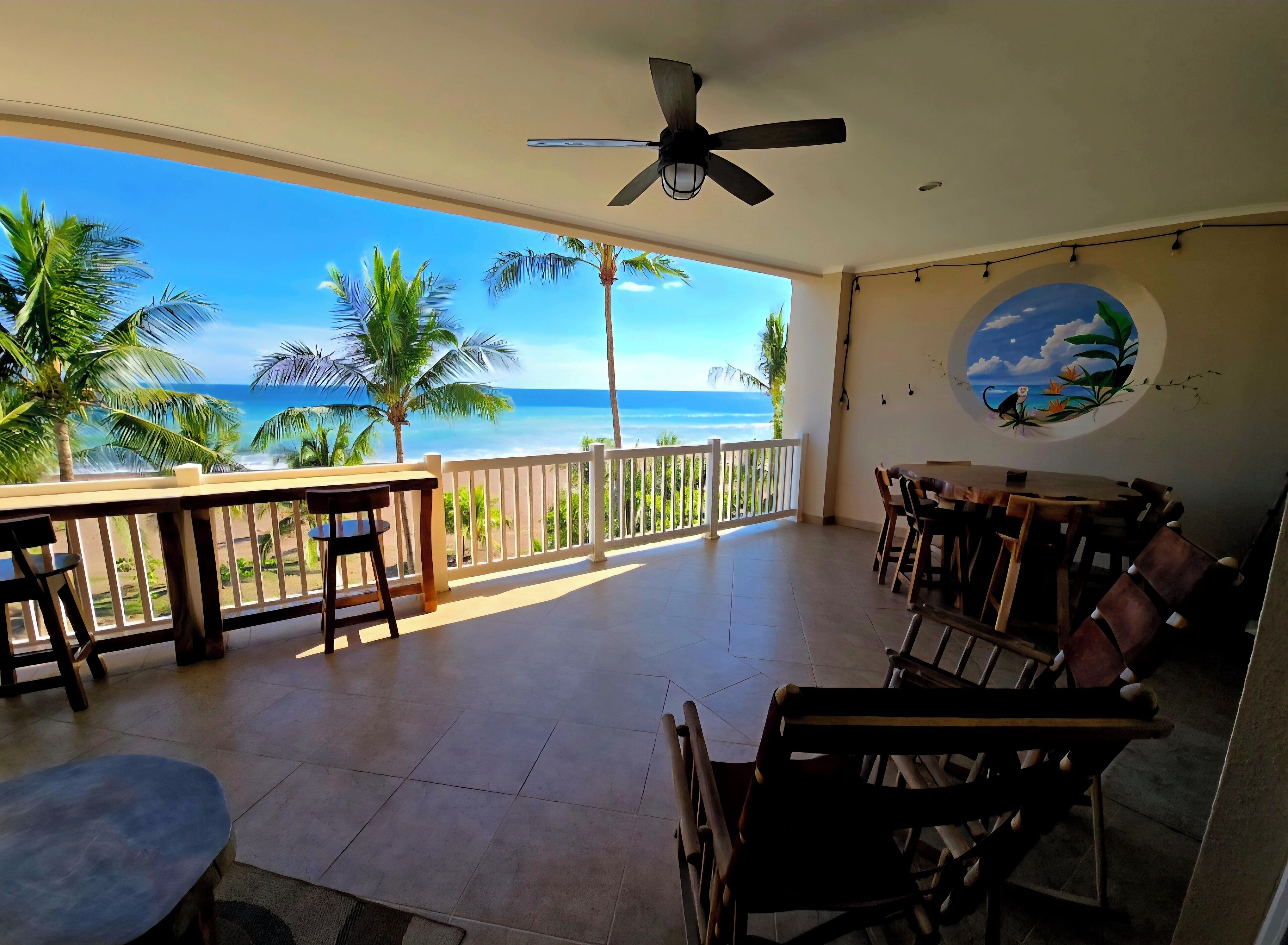 Une pièce avec vue sur l’océan, un ventilateur de plafond, un coin repas avec chaises et tables, et un mur décoratif.