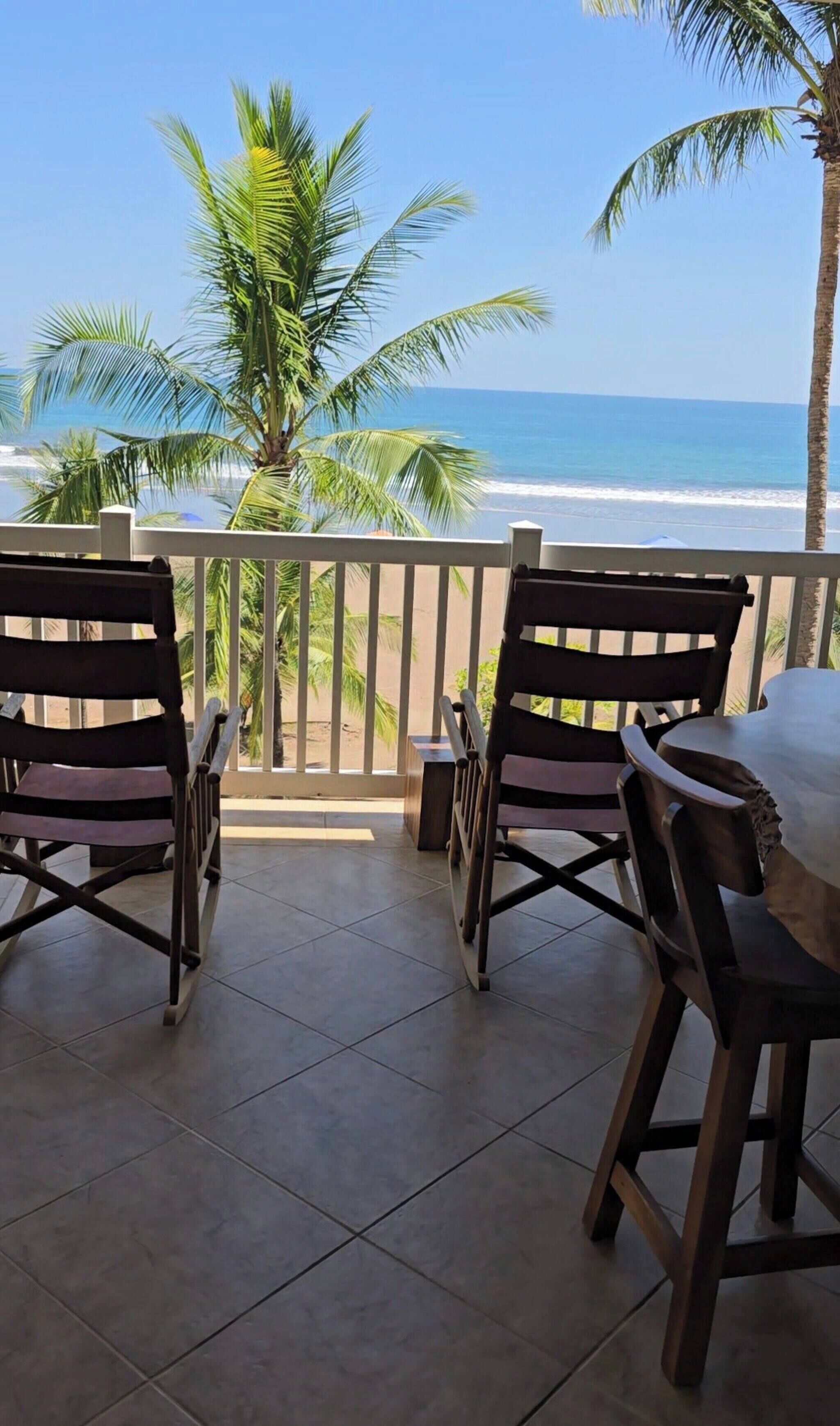 A balcony with two wooden chairs and a small table, overlooking a beach with palm trees and the ocean.