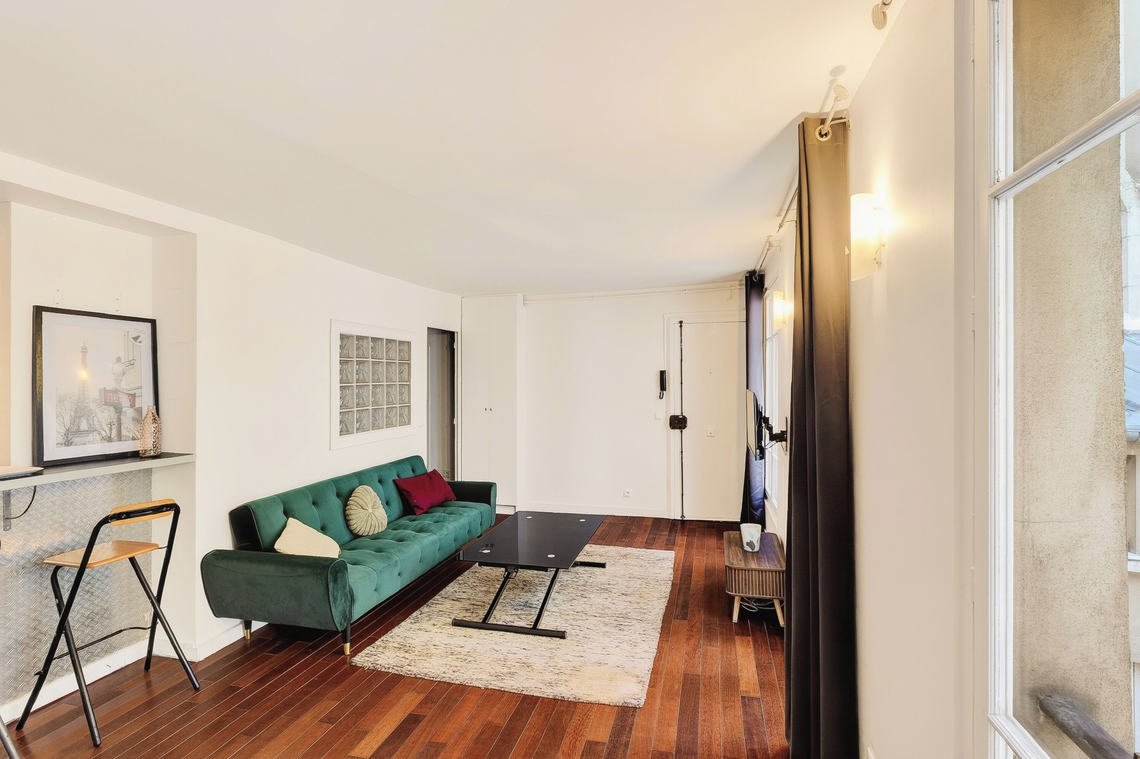 Un salón moderno con un sofá verde, una mesita y una silla. Hay un cuadro en la pared y una puerta que da a otra habitación.