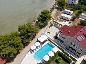 An aerial view of a coastal property with a swimming pool, lounge chairs, and a red-roofed building. - Two Bedroom Sea View Apartment Marisol (Mrljane)