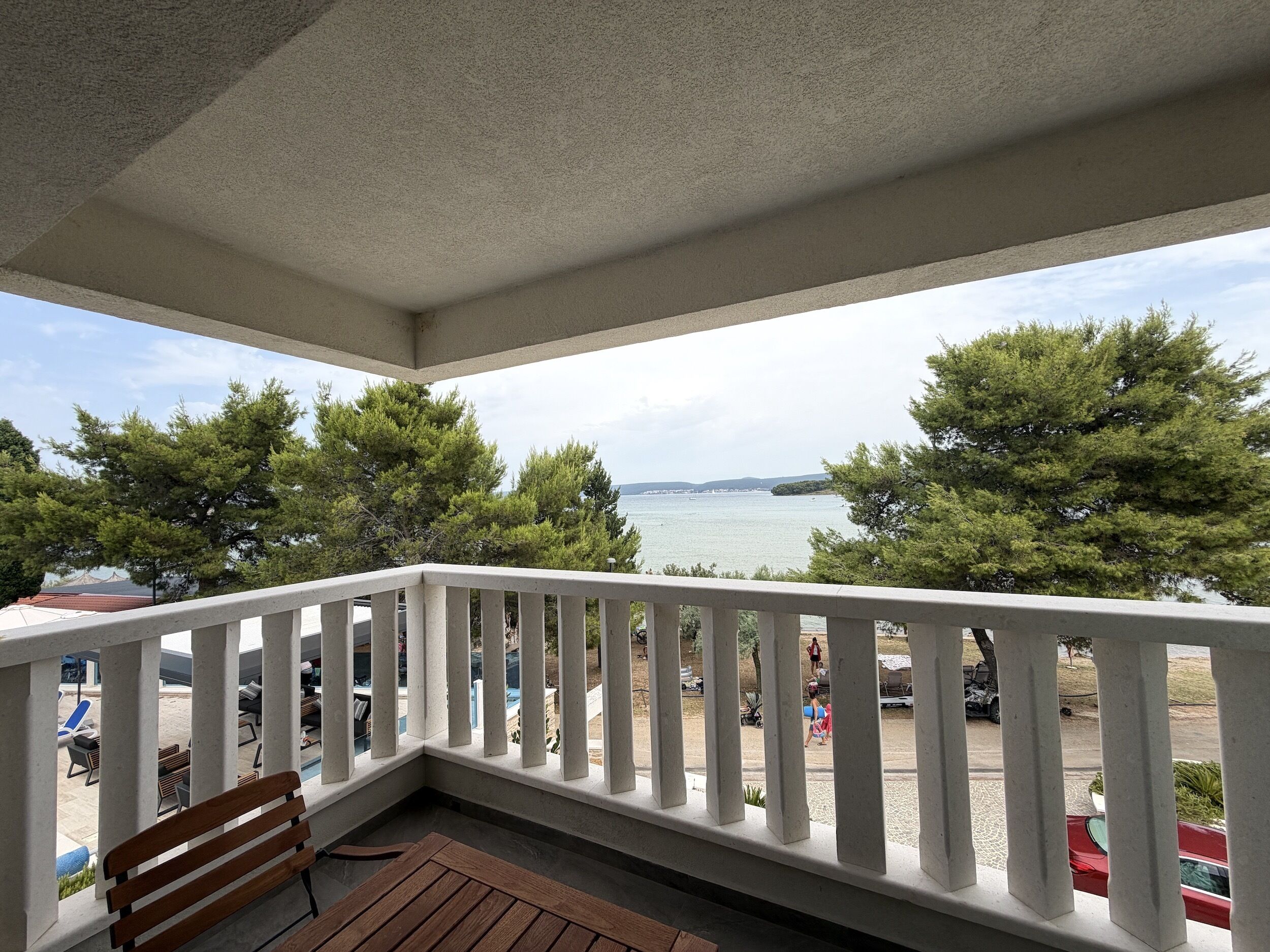 A balcony with a wooden bench, white railing, and a view of the sea and trees.