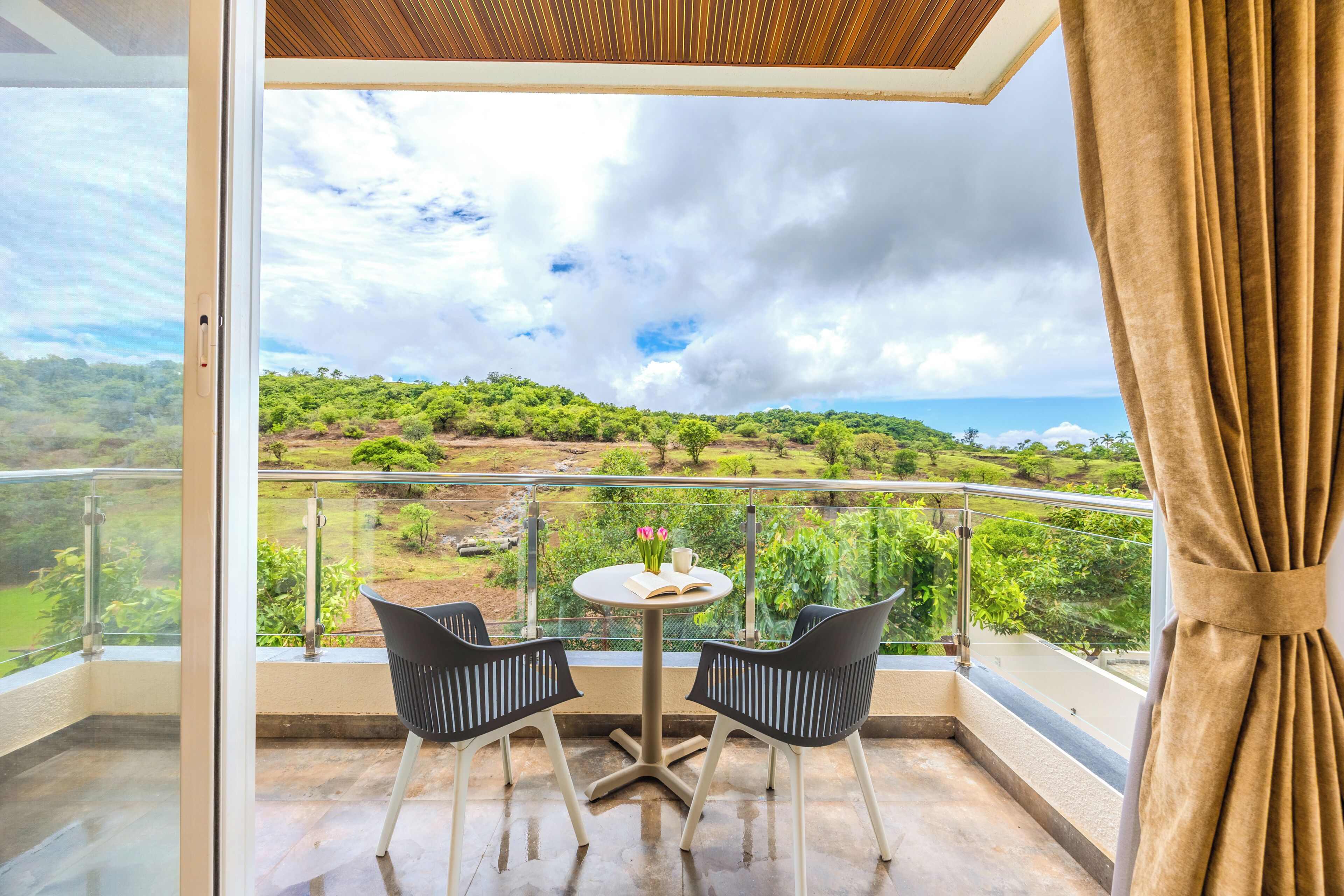 A balcony with a table and chairs, overlooking a lush landscape with trees and a distant view of the sea.