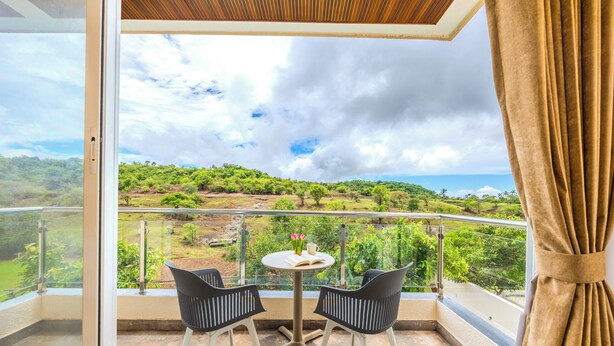 A balcony with a table and two chairs, overlooking a green landscape with trees and a distant view of the sea. - ELIVAAS RK Villa 4BHK Hideaway with Pool (Lonavala)