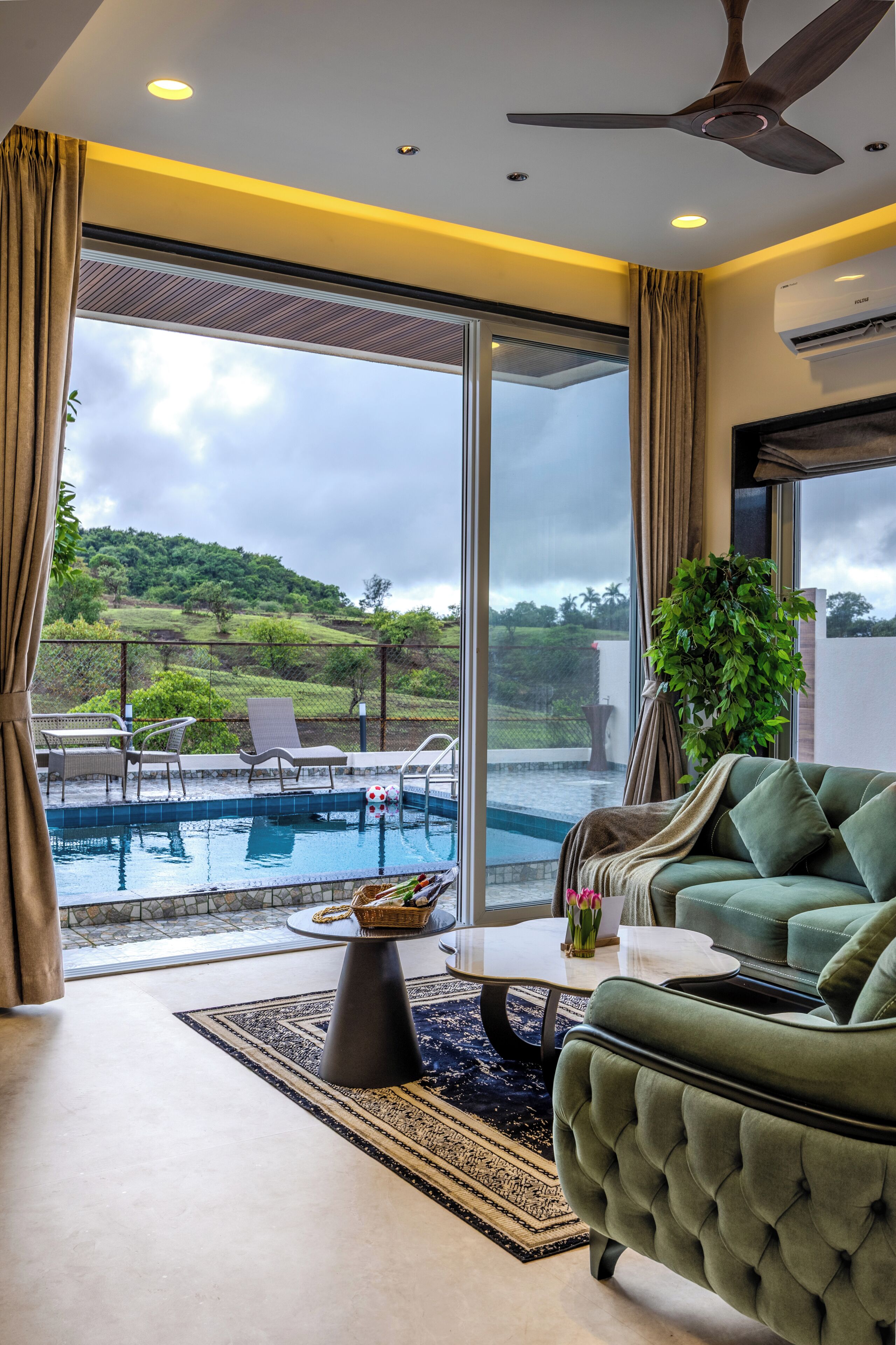 Ett modernt vardagsrum med en grön soffa, ett soffbord och utsikt över en swimmingpool och grönområden utanför.