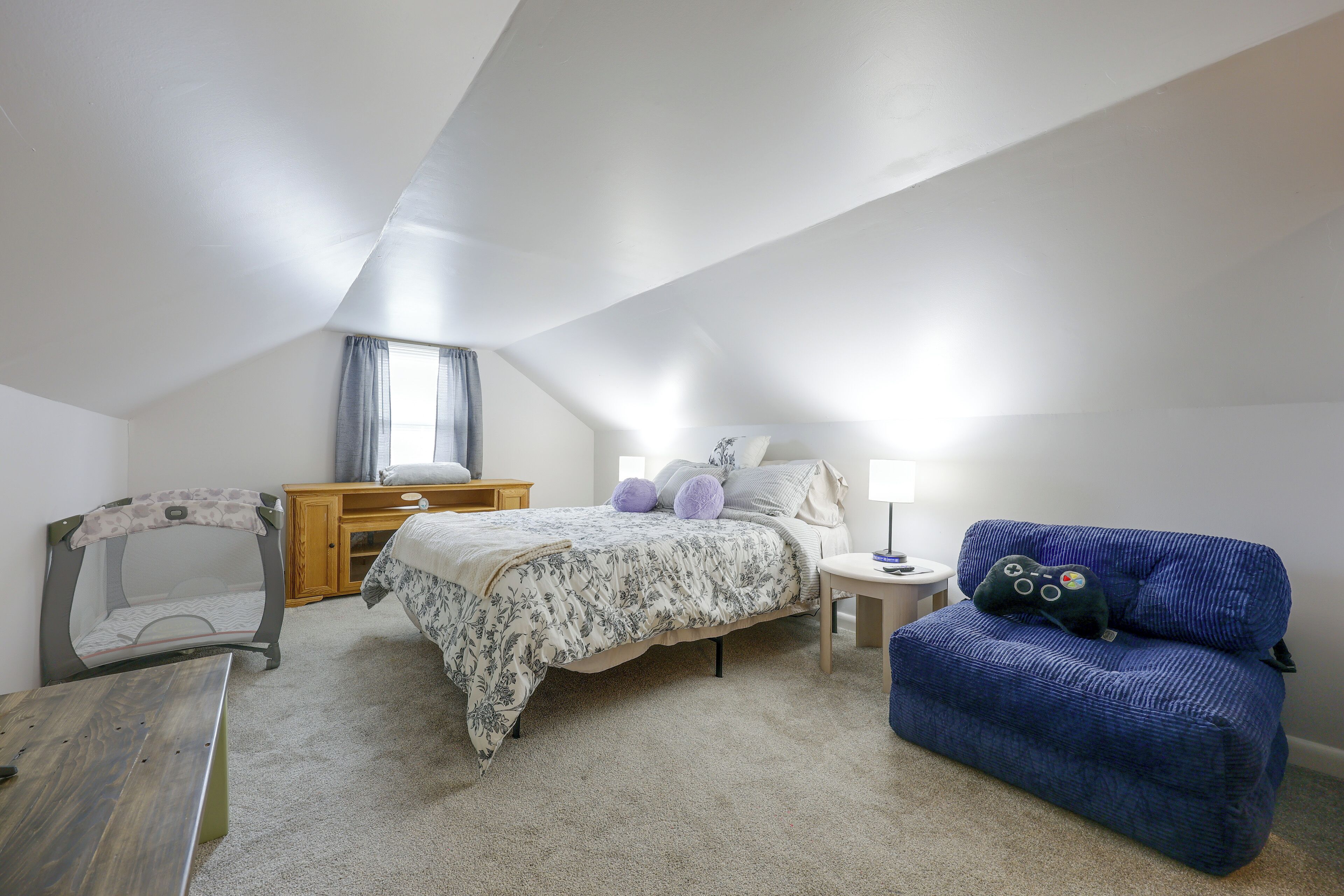 A bedroom with a sloped ceiling, a bed with floral bedding, a blue armchair, a wooden dresser, and a small table with a lamp.