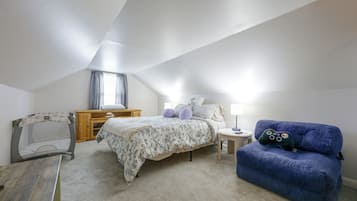 Une chambre avec un plafond en pente, un lit avec une literie à motifs floraux, un fauteuil bleu, une commode en bois et une petite table avec une lampe.