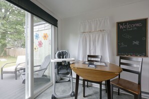 A room with a table, chairs, a baby stroller, a window with a view of trees, and a chalkboard with writing.