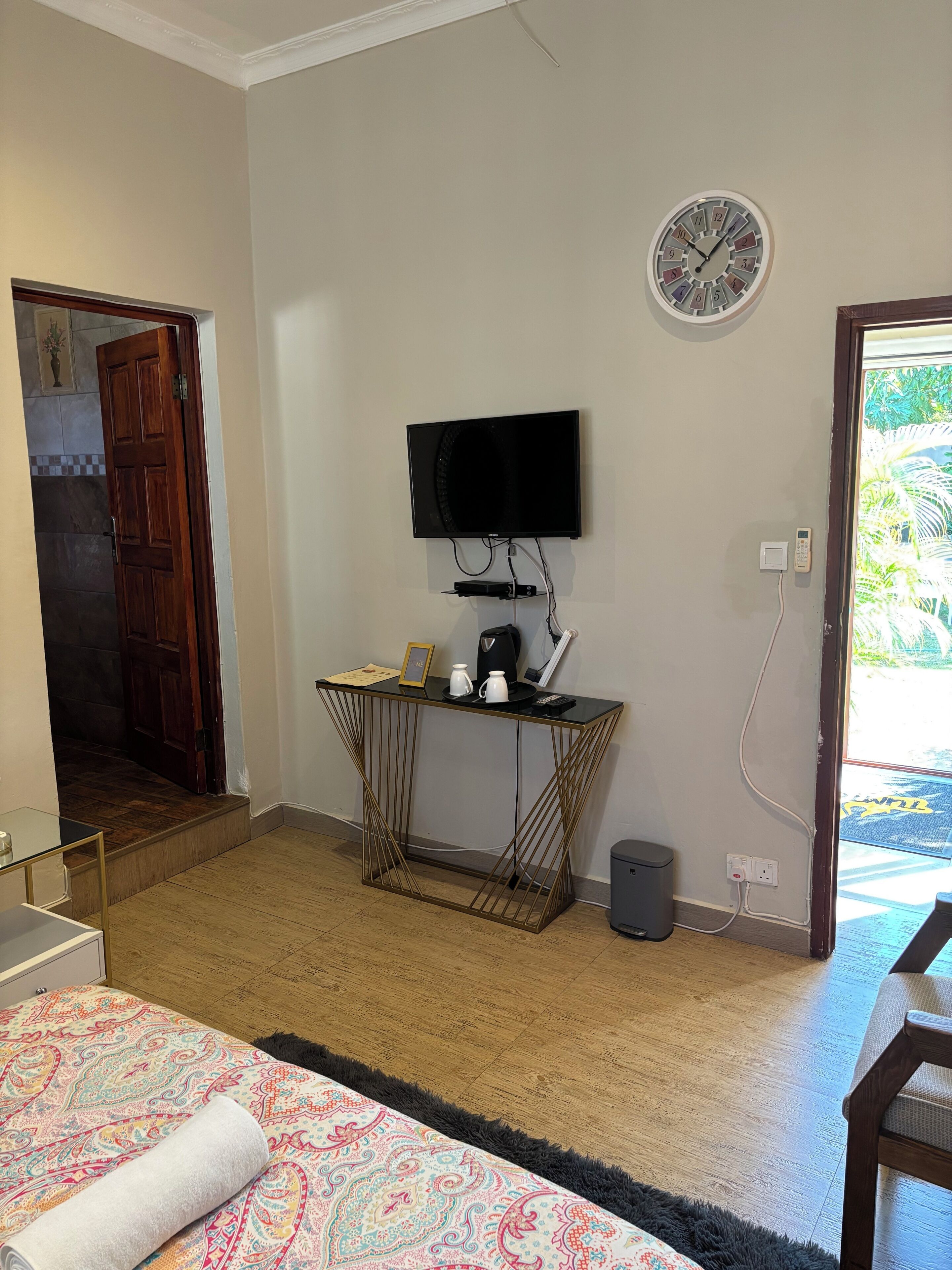 A hotel room with a bed, a TV mounted on the wall, a small table with a kettle and a picture frame, and a door leading to a balcony.