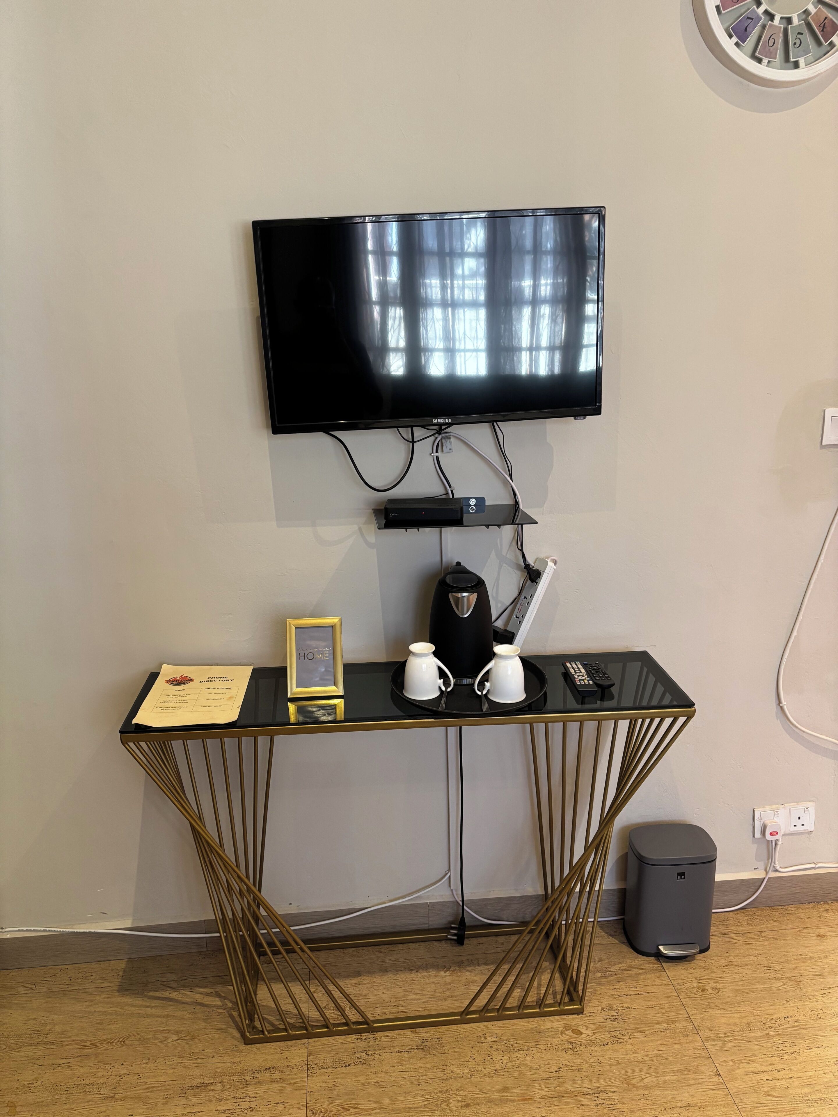 Una habitación con televisor de pared, cafetera, dos tazas, control remoto y un documento enmarcado sobre una mesa negra.