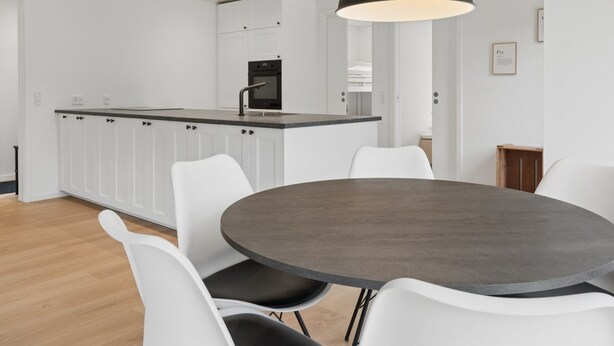 Una cocina moderna con armarios blancos y una isla central, un comedor con una mesa redonda y sillas blancas, y una lámpara colgante arriba.