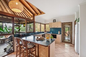 A kitchen with a large island, wooden bar stools, a hanging light fixture, and a view of greenery outside. - River’esque | Spa Bath | Forest Views (Margaret River)