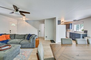 Een moderne woonkamer met een grijze modulaire bank, een eettafel met stoelen, een plafondventilator en een keuken met een raam.