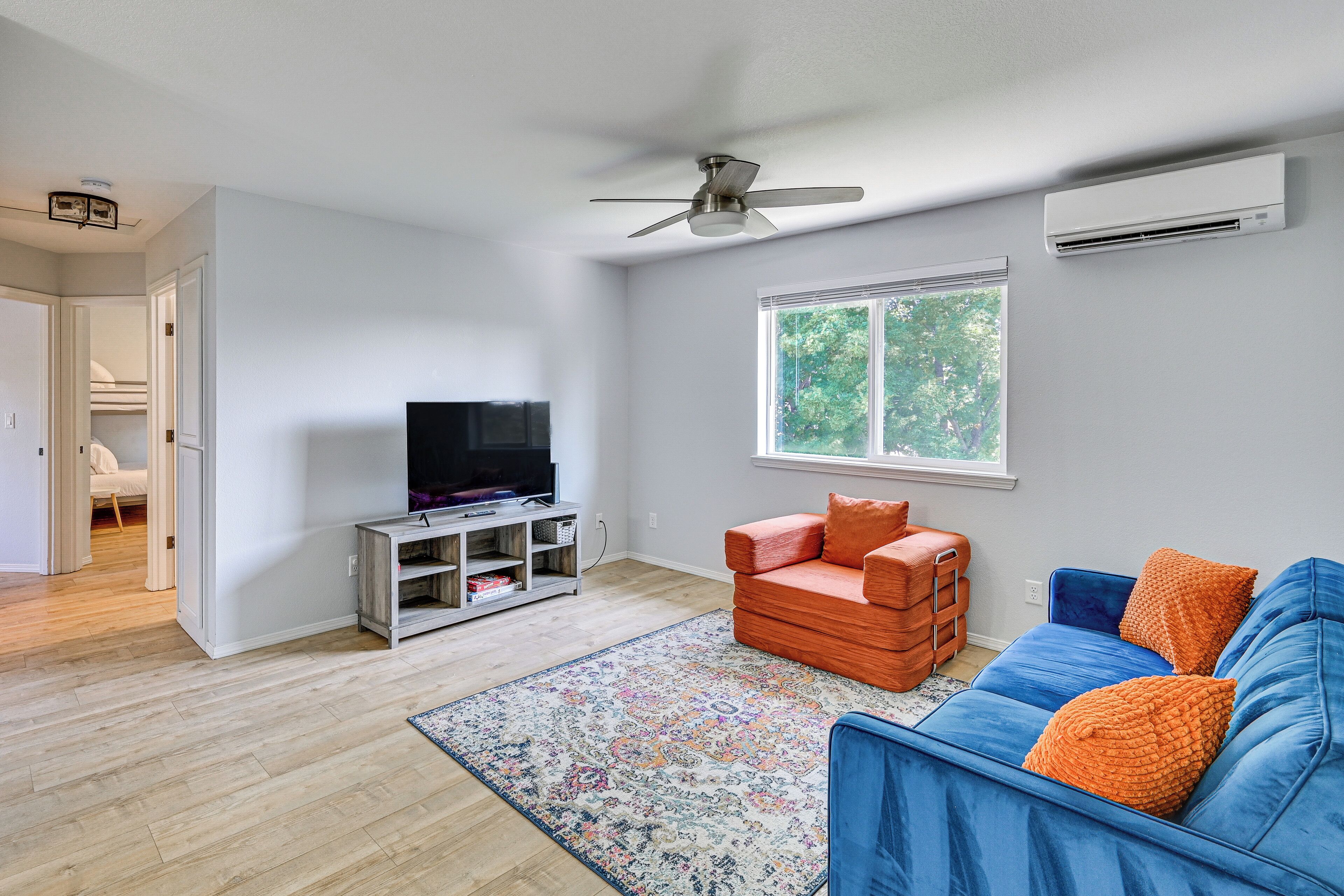 A living room with a television, a blue sofa, an orange chair, a patterned rug, and a window with a view of trees.