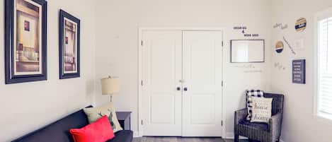 Una sala de estar con un sofá negro, una almohada roja, un tapete estampado, un sillón de mimbre y una puerta blanca.