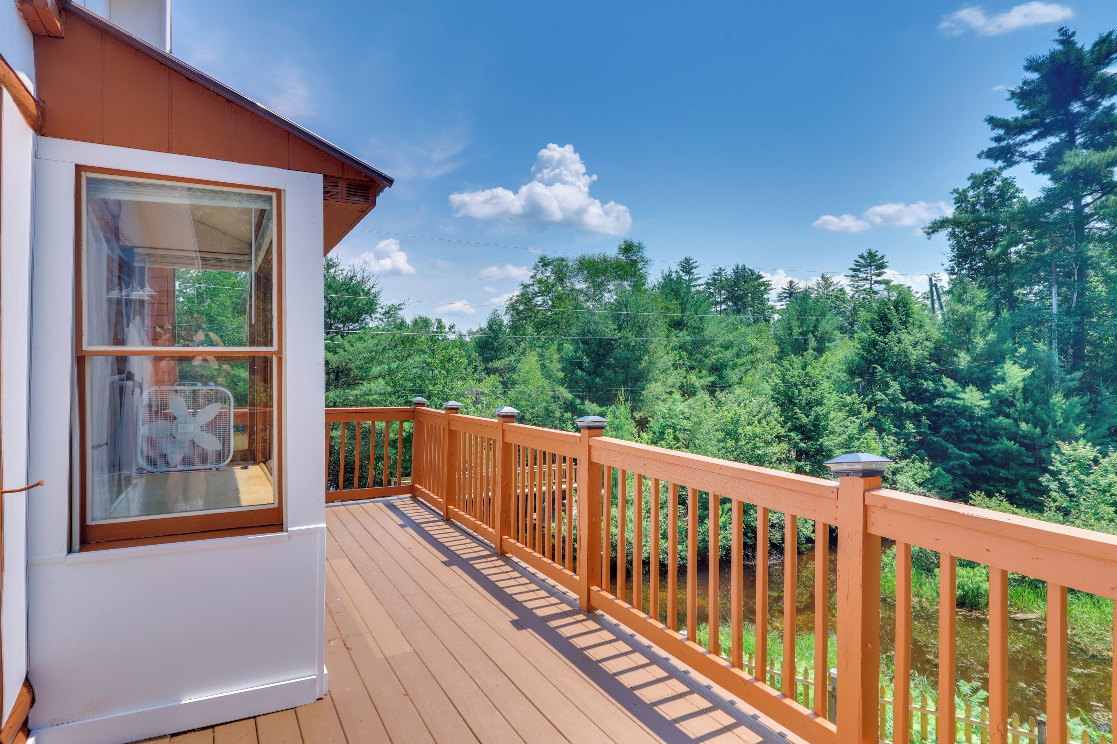 A wooden deck with a railing, a window with a fan, and a view of a forest.