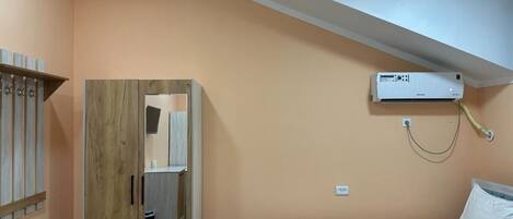 A bedroom with a bed, an air conditioning unit, a wooden wardrobe, and a small bathroom.