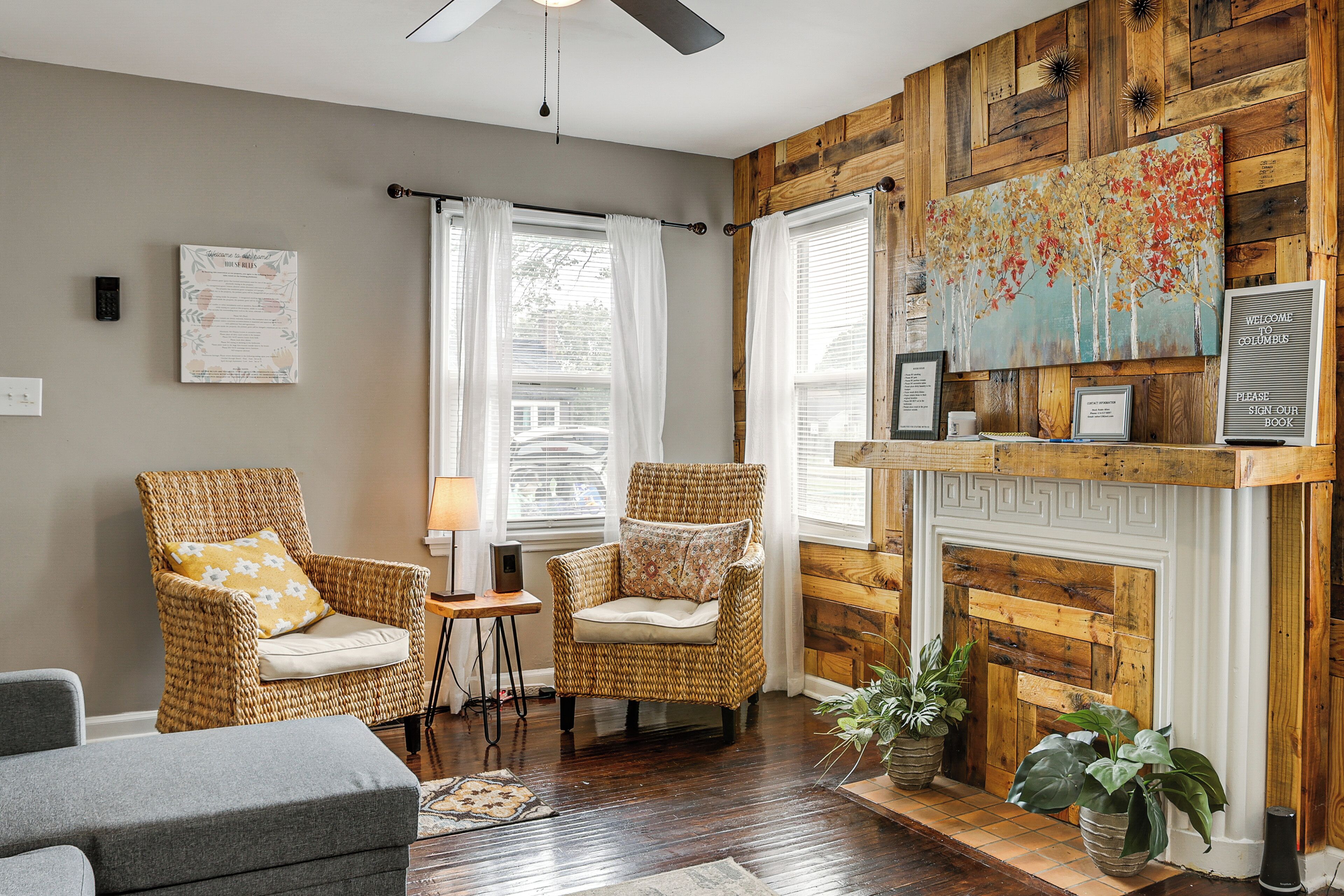 A room with two wicker chairs, a grey sofa, a fireplace with a mantel, and a wooden wall.