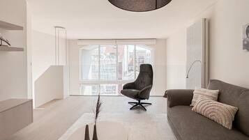 Een moderne woonkamer met een groot raam, een grijze bank, een zwarte fauteuil en een ronde salontafel.