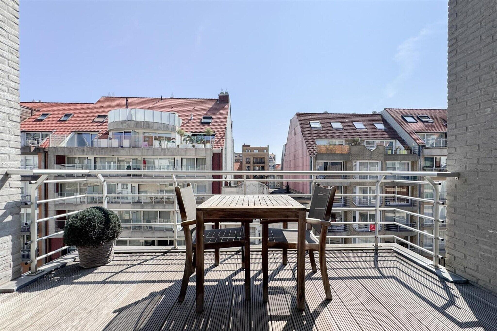 A balcony with a table and chairs, overlooking a residential area with red-tiled roofs.